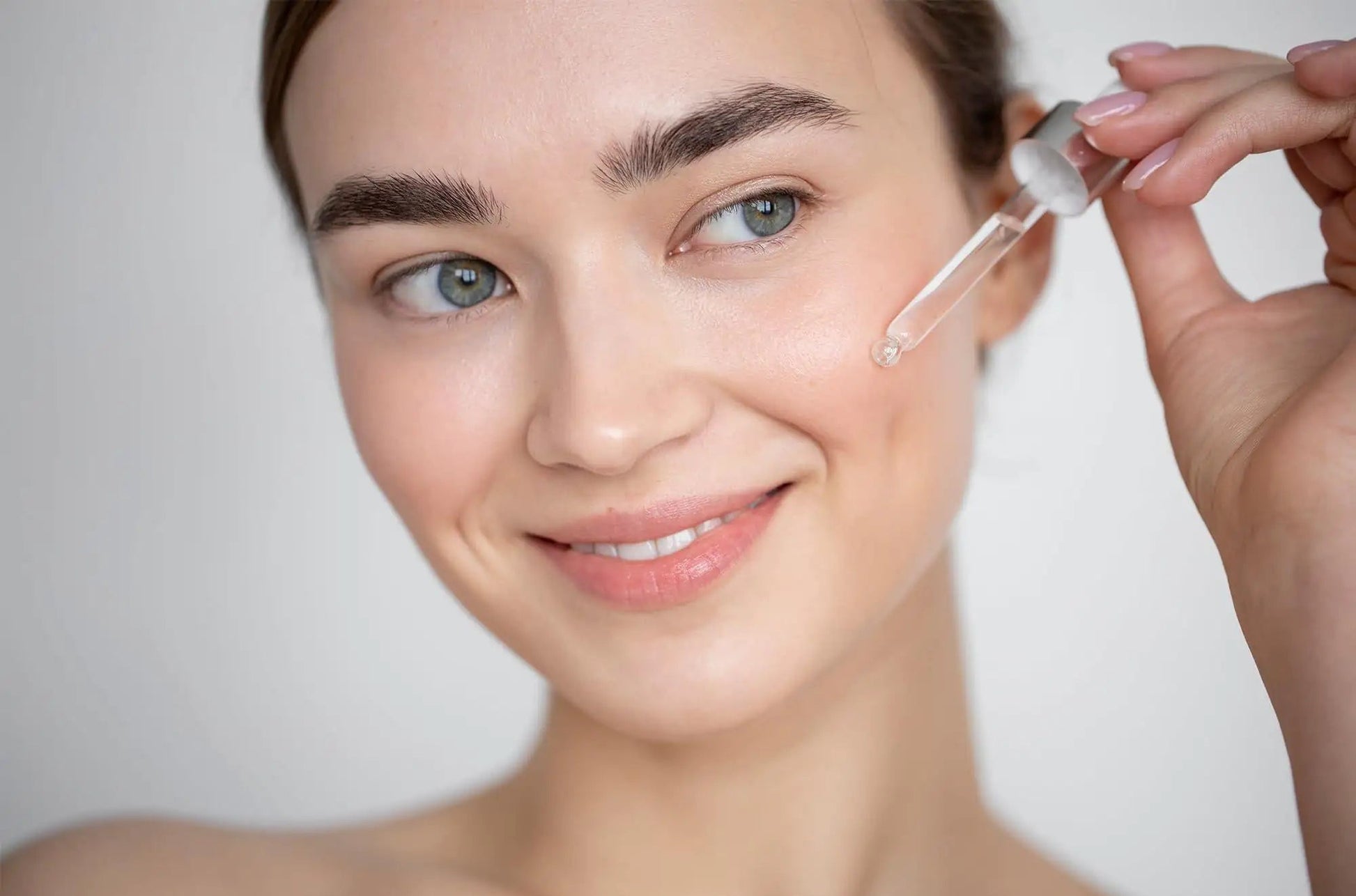 Woman applying skincare serum to her face with a dropper, against a neutral background.