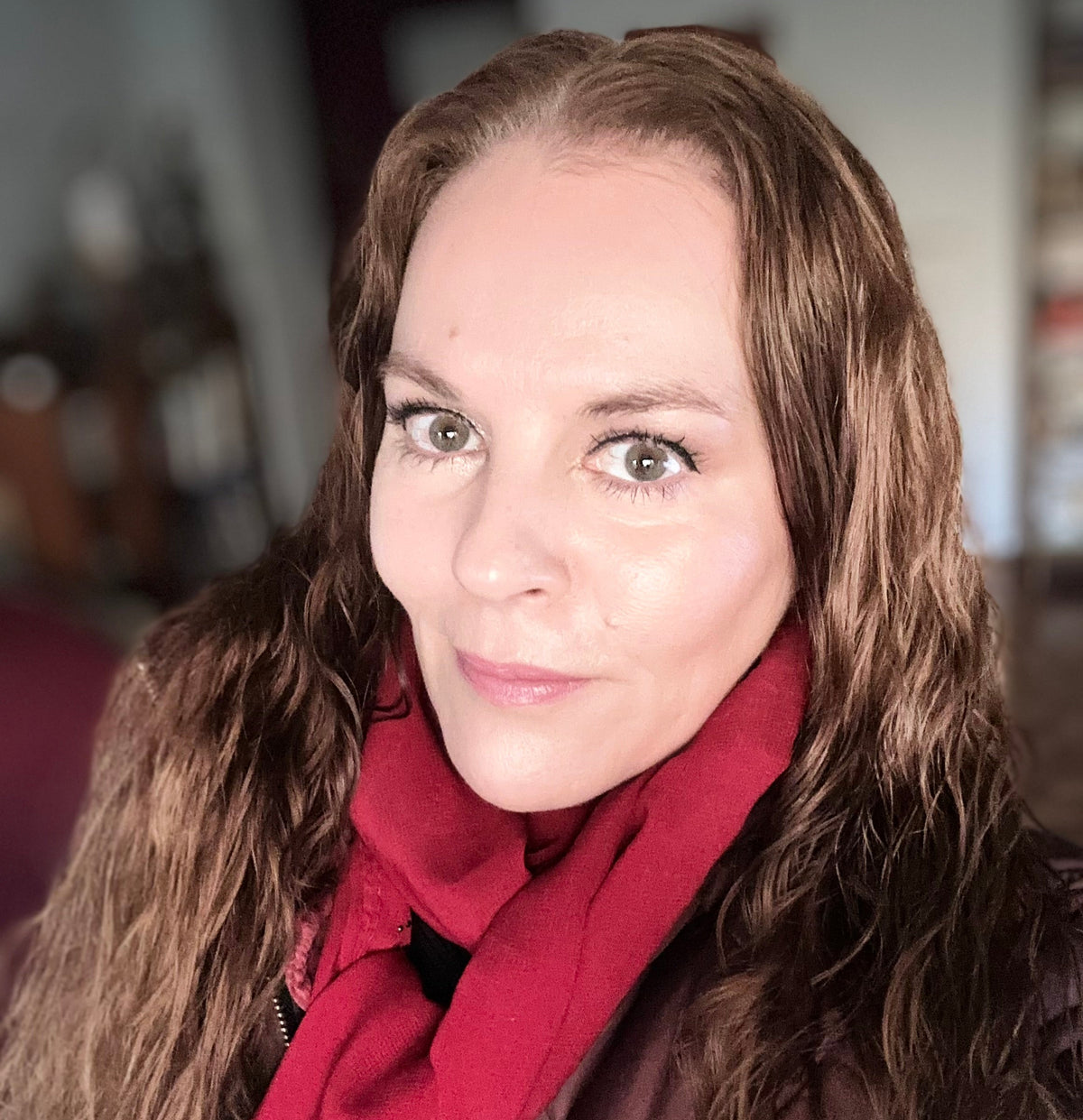 A close-up portrait of a woman with long brown hair wearing a bright red scarf and a dark quilted jacket, set against a softly blurred indoor background.