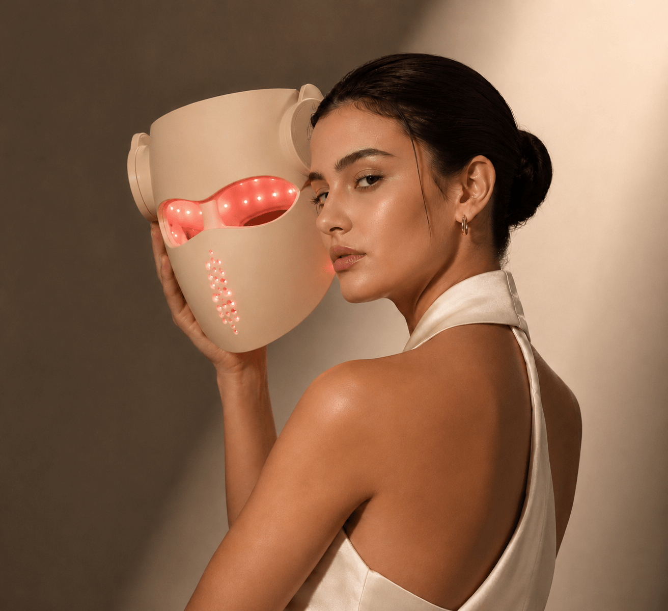 Woman holding a beige LED face mask with visible lights against a neutral background
