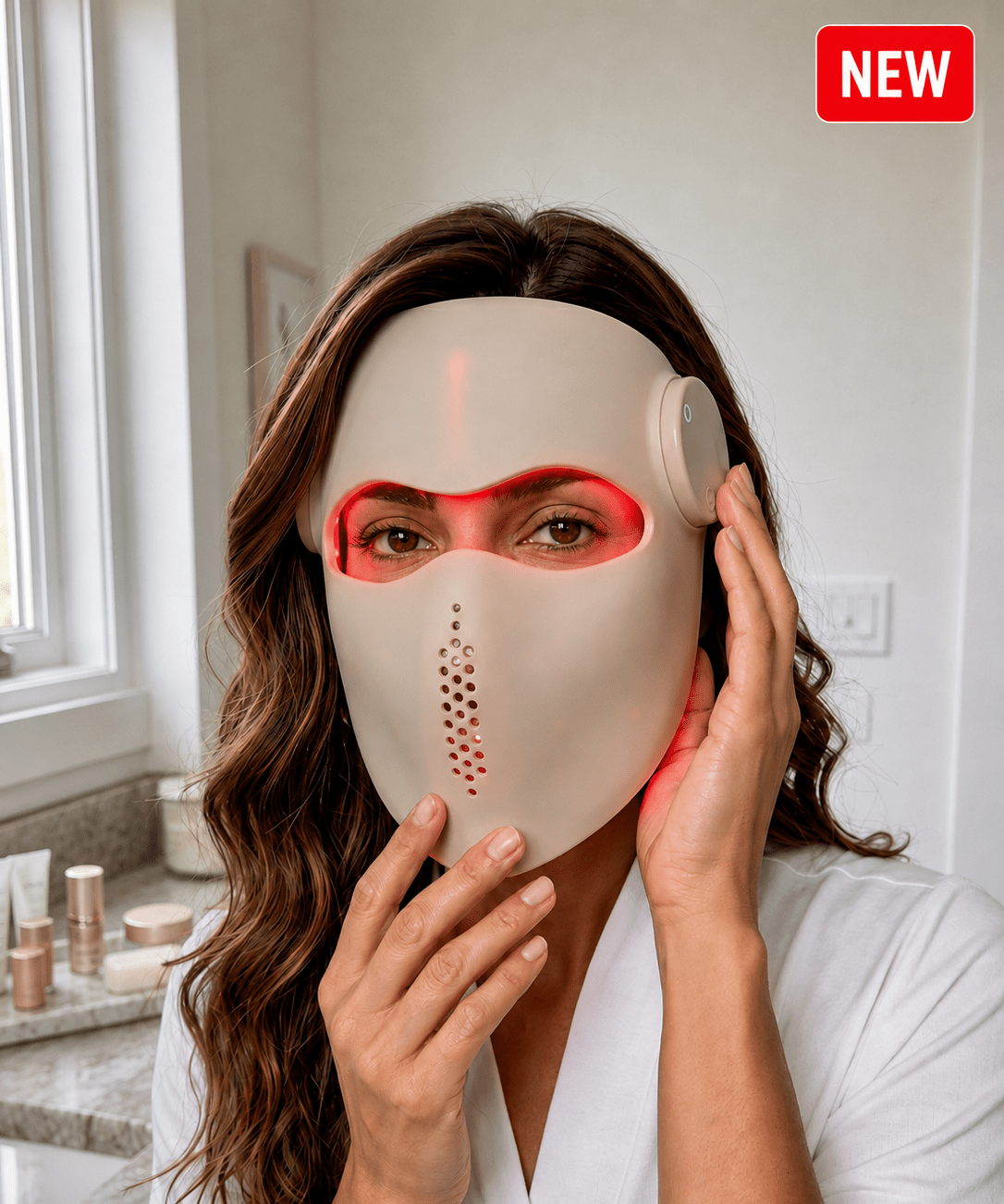A woman wearing a beige LED mask with red light glowing from the eyes, nose, and cheek, in a bright bathroom setting.