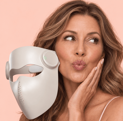 A woman in a pink satin camisole holds a white LED facial mask up next to her face against a solid light pink background.