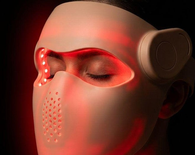 A person wearing a white LED light therapy face mask with red illuminated lights, shown in a close-up profile against a black background.