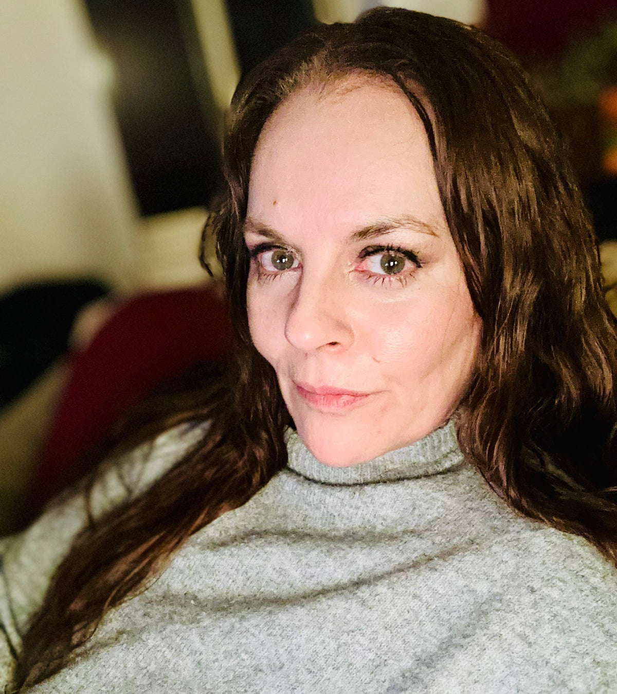 A close-up portrait of a woman with long brown hair wearing a light gray turtleneck sweater, looking directly at the camera in an indoor setting.