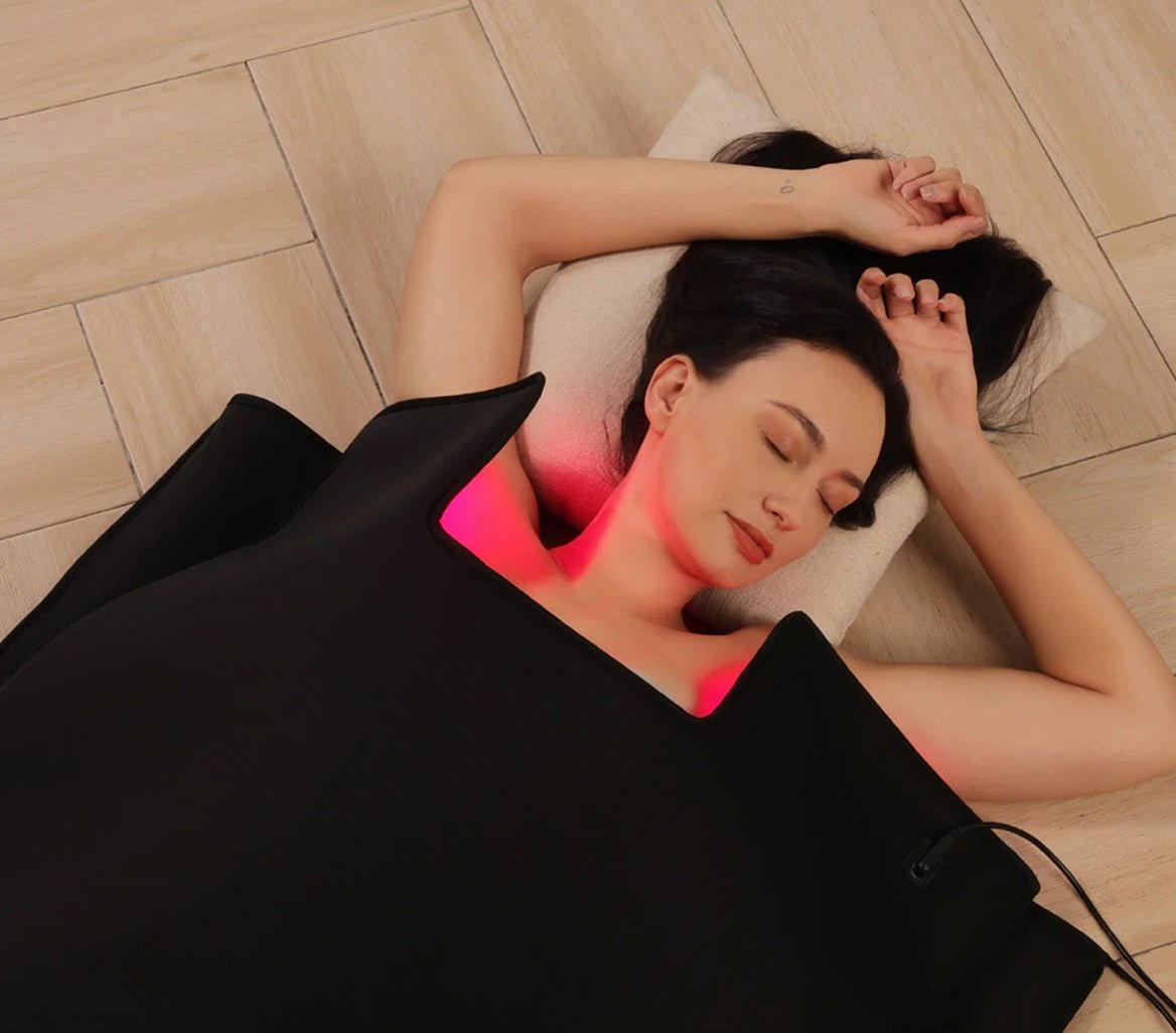 Woman lying on a Sauna Blanket with a glowing red light on her neck, on a wooden floor.