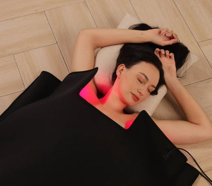 Woman lying on a Sauna Blanket with a glowing red light on her neck, on a wooden floor.
