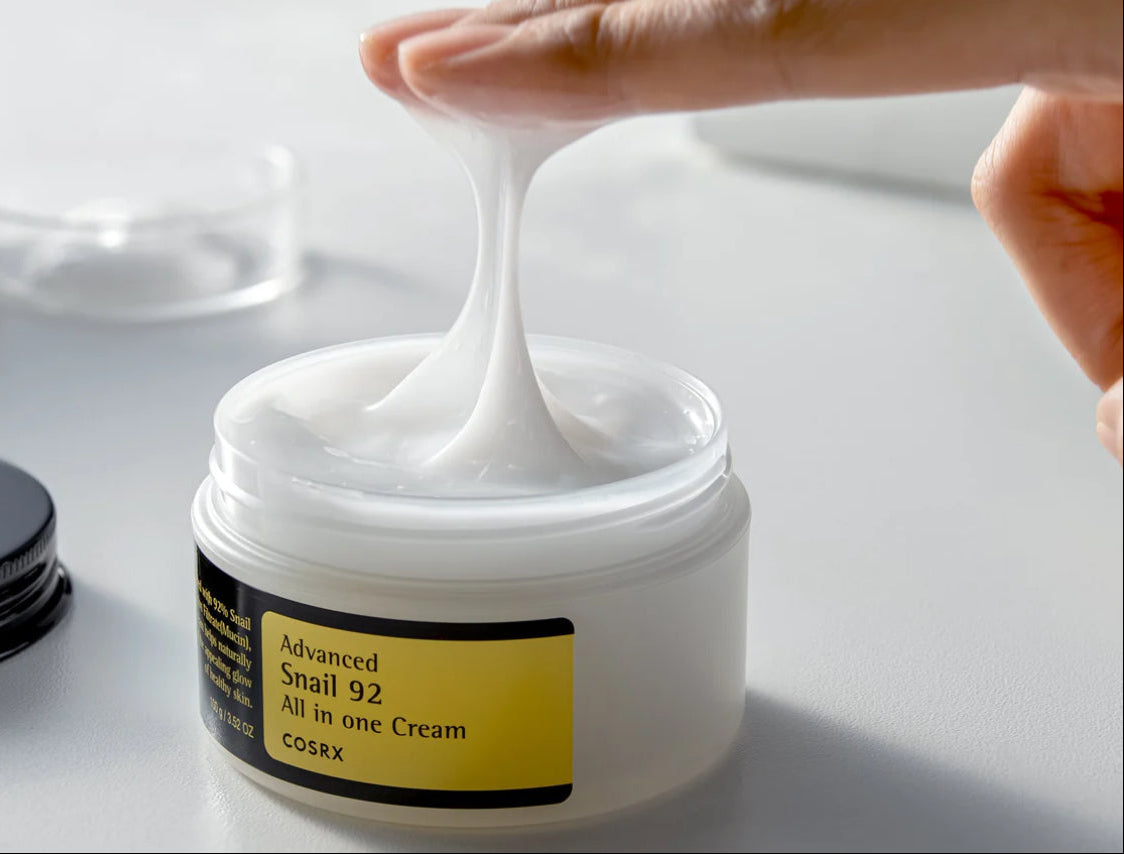 Hand applying cream from a jar labeled 'Advanced Snail 92 All in One Cream' on a white surface.
