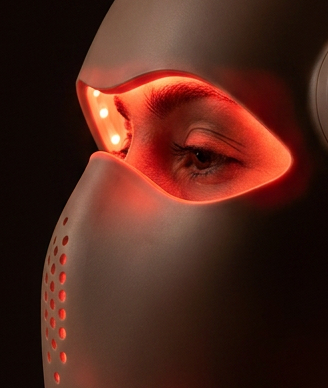 A person in profile wearing a futuristic LED light therapy mask that glows red around the eyes and nose area, set against a dark background.